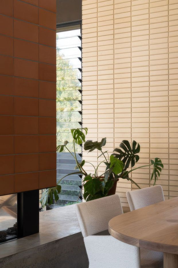 A refined dining area with a round light timber dining table and upholstered cream dining chairs. The room features a dramatic vertical composition of warm terracotta wall tiles beside pale textured brick-like wall cladding, a tall louvred window, a dark structural fireplace or column element, and indoor tropical foliage. The overall design is warm, contemporary, and material-focused with abundant natural light.
