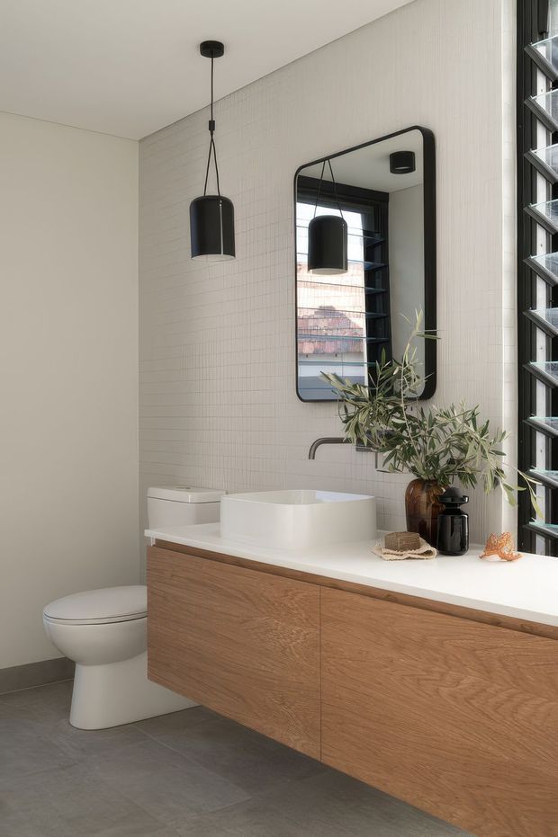 A contemporary bathroom with a floating natural timber vanity, white solid-surface benchtop, and round above-counter basin. The walls are clad in small vertical white tiles, with large grey floor tiles, a wall-mounted black-framed mirror, black pendant light, and black louvred window. A toilet and restrained botanical styling complete the calm, refined space.