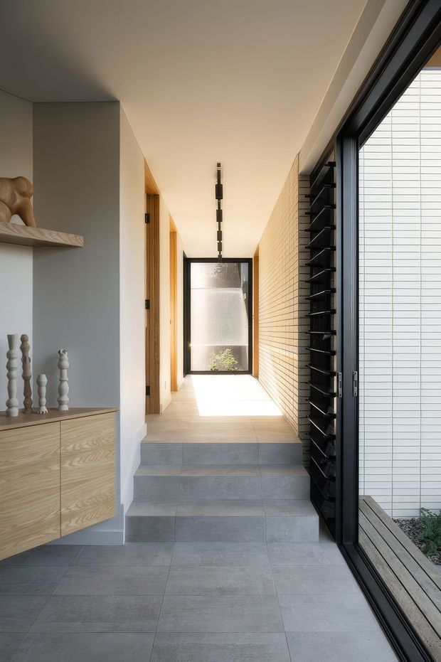 A bright, narrow residential hallway with a short flight of grey tiled steps leading to a sunlit passage and glazed door. The space features a full-height black-framed glazed opening with horizontal louvres on the right, a pale textured brick wall, warm timber door frames and a built-in light timber console with decorative objects on the left. The design is clean-lined and contemporary, with strong natural light and a calm neutral palette.