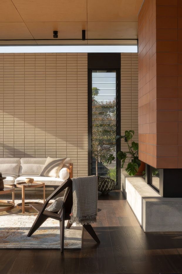 A warmly lit contemporary living room with a low cream sofa, timber coffee tables, an armchair, and a large neutral rug. The room features dark timber flooring, extensive narrow-format terracotta-toned tiled walls, a timber-lined ceiling, black-framed vertical glazing, and a built-in concrete and tiled architectural feature along the right side. Indoor planting and strong afternoon sunlight create a calm, refined atmosphere.