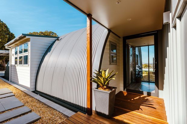 Exterior view of a compact contemporary dwelling with a distinctive curved corrugated metal roof and matching light cladding. The entrance is approached via a timber deck and stepping-stone path, with large glazed doors and windows opening toward a bright view beyond. A potted plant sits beside the sheltered entry.