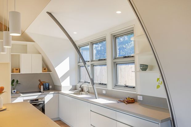 Bright contemporary kitchen beneath a sloped ceiling, with streamlined white cabinetry, a light grey benchtop and splashback, integrated appliances, and three large timber-framed windows. A sculptural curved structural element and pendant lights add visual interest, while sunlight creates a warm, airy atmosphere.