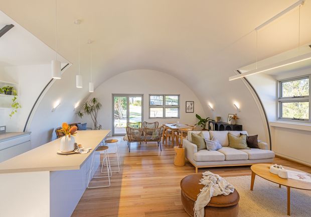 Open-plan, light-filled living and dining space with a contemporary kitchen along the left wall. The room has a distinctive curved, barrel-vaulted ceiling and matching curved side walls, warm timber flooring, pale built-in cabinetry, a light sectional sofa, timber dining furniture, and large windows providing views to greenery outside. Linear pendant and track lighting emphasize the elongated vaulted form.