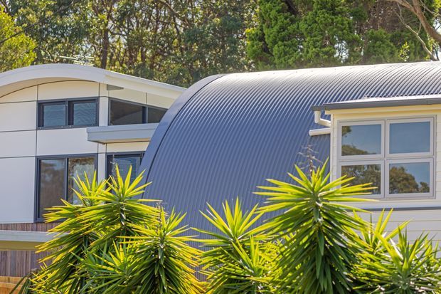 Exterior view of a contemporary residential building with a distinctive curved corrugated metal roof and a pale, modern facade. Dark-framed glazing is visible on the left volume, while the right section has white-framed multi-pane windows. Layered yucca-like landscaping partially obscures the lower facade, with mature trees behind the building.