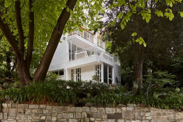 A multi-storey white residential building set within dense mature garden landscaping. The facade features stacked open balconies with white metal balustrades, large glazed openings, slender structural posts, and a flat roofline. A rough stone retaining wall and abundant established planting frame the foreground, giving the property a secluded, leafy character.