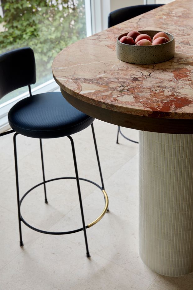 Close-up interior view of a refined kitchen or dining area featuring a round, thick natural-stone tabletop with prominent pink, cream and rust veining, supported by a dark timber apron and a fluted pale cylindrical pedestal. Two black upholstered bar stools with slim black metal legs and circular footrests sit beside the table, one with a brass-gold footrest detail. Large glazing overlooks greenery, creating a bright, elegant setting.