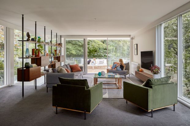 Bright, spacious contemporary living room with floor-to-ceiling glazing on multiple sides, opening to an outdoor terrace. The room features a large grey sofa, green upholstered lounge chairs, a timber-and-glass coffee table, an open black-framed display shelving unit, a wall-mounted television, and a neutral carpeted floor. Abundant greenery is visible outside, creating an airy connection to the landscape.