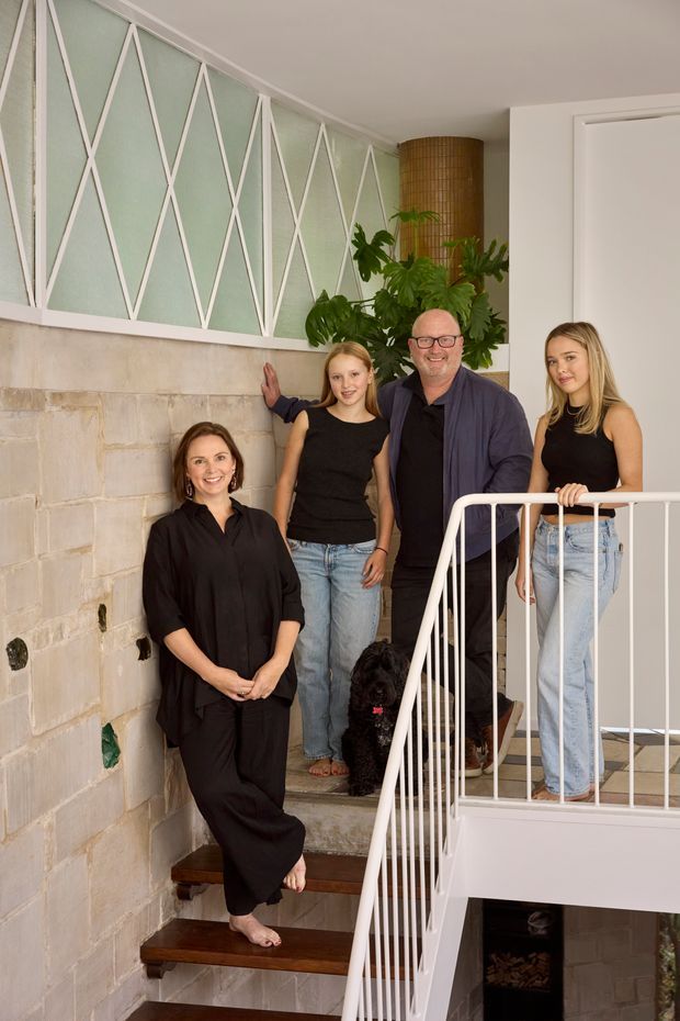 A vertical interior photograph of a residential stairway with a family gathered on the steps and landing. The stairwell features dark timber treads, white painted balustrades, pale masonry or concrete-block walls, and a striking pale green geometric lattice wall panel with white diagonal trim. A large indoor plant and cylindrical woven pendant light add warmth to the space.