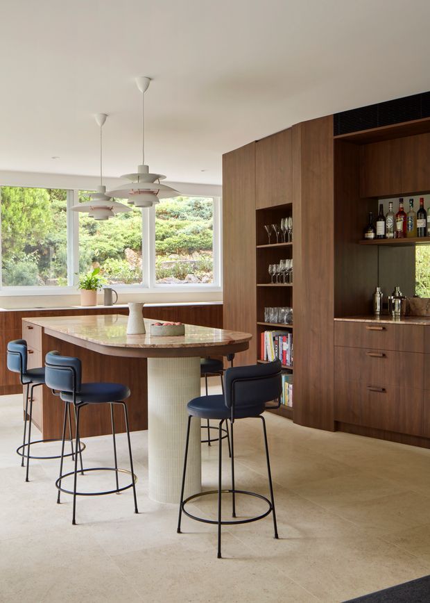 A bright, contemporary kitchen and home bar area with extensive dark timber cabinetry, an integrated island with a rounded stone benchtop and cylindrical textured pedestal, and several navy upholstered bar stools. The kitchen opens toward large windows overlooking greenery. Pendant lights hang above the island, while open shelving displays glassware and bottles. The space has a warm neutral palette, polished stone or concrete-like flooring, and a refined mid-century-influenced furnishing scheme.