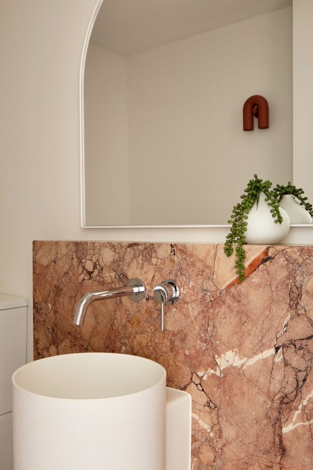 Close-up view of a refined bathroom vanity featuring a white vessel basin, wall-mounted chrome tapware, and a dramatic pink-brown veined stone backsplash and counter. A large arched mirror with a slim light frame occupies the wall, complemented by a small red sculptural wall ornament and trailing greenery in white vessels. The space has a warm, elegant, contemporary feel.