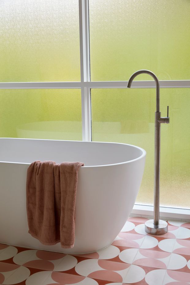 A bright bathroom featuring a freestanding white oval bathtub, a tall brushed-metal floor-mounted bath filler, and a warm terracotta towel draped over the tub. Large frosted window panels provide diffused green-tinted light, while the floor has a bold repeating pink, terracotta, and white geometric tile pattern.