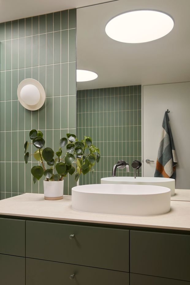 A contemporary bathroom vanity featuring a round white vessel basin on a pale stone countertop, dark green built-in cabinetry, and full-height muted sage-green rectangular wall tiles with light grout. A wall-mounted circular light, chrome tapware, potted plant, mirror, and striped towel complete the calm, modern interior.
