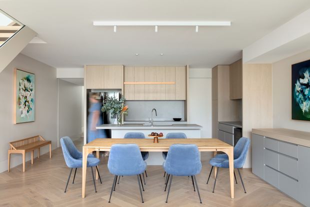 Bright contemporary open-plan kitchen and dining area with a pale timber dining table surrounded by blue upholstered chairs. The kitchen features light wood cabinetry, a grey stone-look island benchtop, integrated appliances, and a simple linear track light. Built-in storage continues along the right wall, while abstract artwork and a timber bench add restrained decorative detail.
