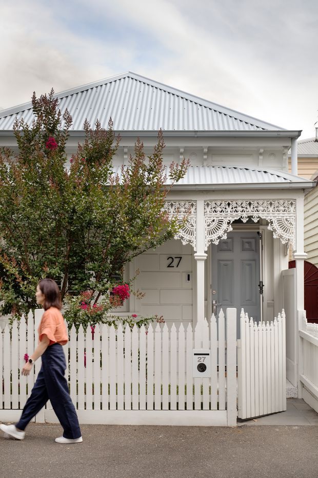 Street-facing view of a compact heritage-style cottage with a white corrugated gable roof, pale weatherboard cladding, ornate decorative lacework over the verandah, white picket fencing, and a grey panelled front door. Flowering shrubs partially frame the facade, with the house number 27 visible beside the entry.