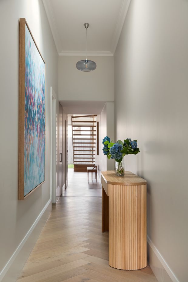 A refined residential hallway viewed toward a bright stair landing. The long narrow corridor has pale greige walls with decorative ceiling cornices, light timber herringbone flooring, white skirting, and a large colourful framed artwork. A curved fluted timber console with a stone or timber-look top sits along the right wall, topped with a glass vase of blue hydrangeas. A contemporary pendant hangs centrally, while a timber open-riser staircase and chair are visible at the far end.