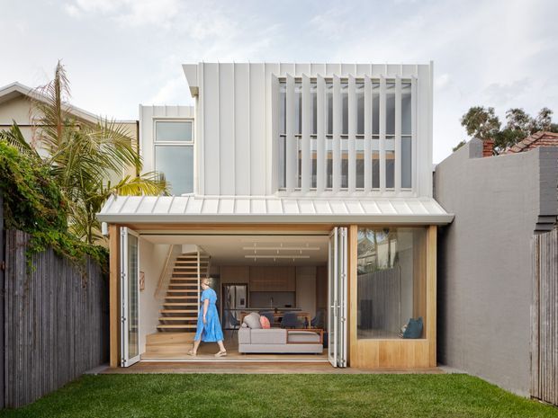 A contemporary two-storey residential extension viewed from the rear garden. The facade combines white vertical cladding and a prominent vertical privacy screen over upper-level glazing, with a light metal skillion roof over a fully glazed ground-floor living area. Sliding or folding glass doors open onto a small lawn, revealing a living room, timber staircase and built-in joinery. Neighbouring urban residential properties, fencing and tropical planting frame the scene.