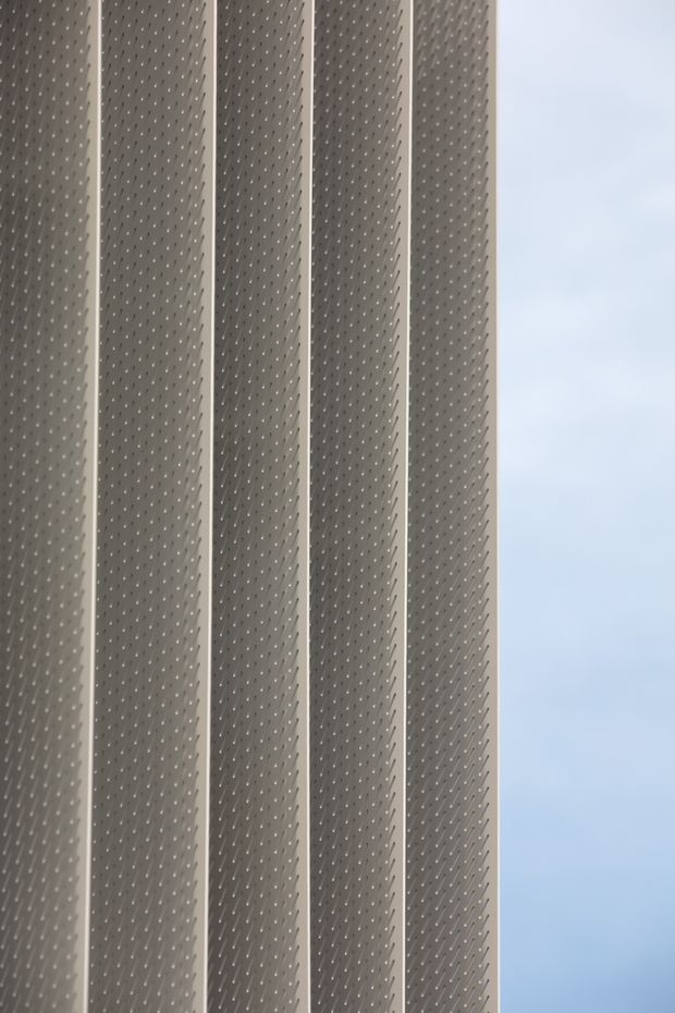 Close-up view of a tall, repetitive facade screen composed of narrow vertical grey perforated panels with lighter structural ribs between them. A pale blue sky is visible along the right edge, indicating an exterior architectural detail. The image is minimalist and strongly pattern-driven, with no interior space or furnishings visible.