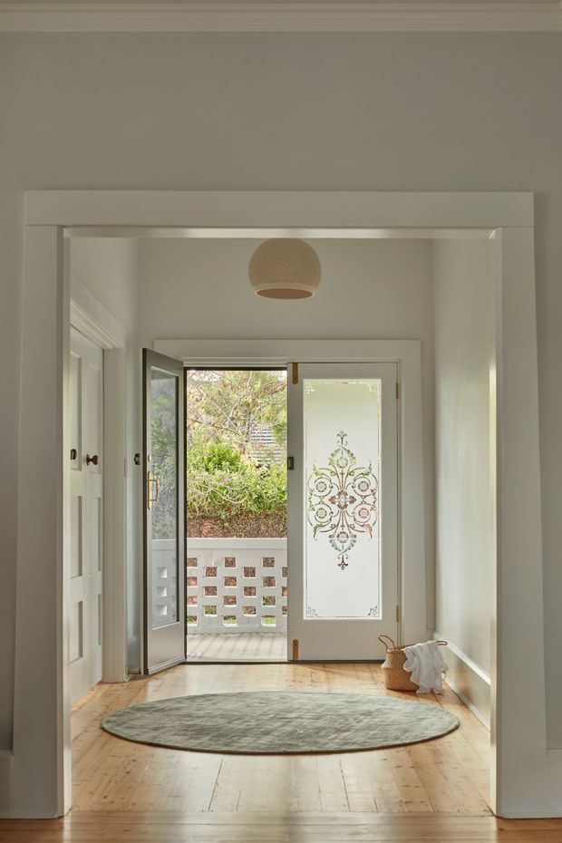 A bright residential entry hall with pale timber floorboards, white trim and panelled doors, and a glazed front door featuring decorative patterned glass. The door opens to a small exterior landing with a perforated brick privacy wall and greenery beyond. A round pendant light, oval rug and woven basket add understated decorative detail.