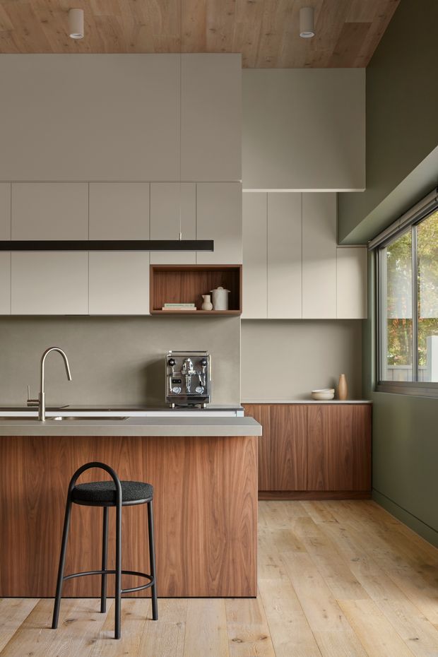 A compact, refined kitchen with warm timber cabinetry and ceiling, muted taupe-grey wall cabinetry and backsplash, and a slim stone island benchtop. A black-framed bar stool sits at the island, while a large window brings in natural light. The kitchen features integrated upper storage, a timber display niche, stainless-steel sink and tapware, and a compact coffee machine.