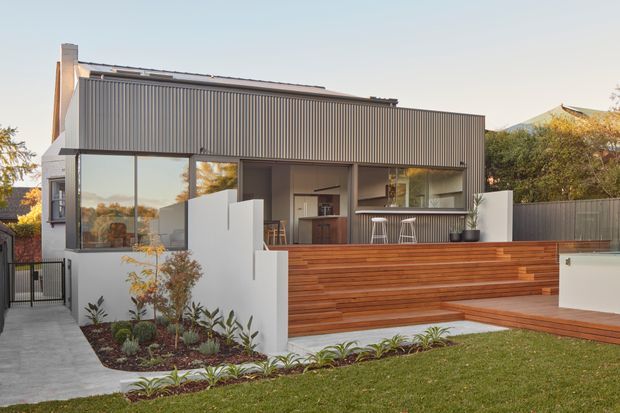 Exterior view of a contemporary two-storey residence with a dark vertically ribbed upper facade, large glazed openings, and an open ground-floor entertaining area. Broad timber-clad steps form a raised terrace, bordered by white rendered walls, simple landscaping, and a lawn. The terrace includes a built-in outdoor kitchen or bar with stools and a covered seating area.