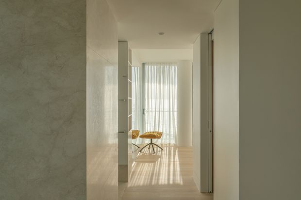 A softly lit minimalist interior hallway or circulation space leading toward a bright sitting area. The scene features smooth off-white walls, pale timber flooring, a tall sheer-curtained window, built-in open shelving, and a small sculptural table with a warm-toned chair or stool. Strong filtered sunlight creates vertical shadows across the floor, giving the space a calm, airy atmosphere.