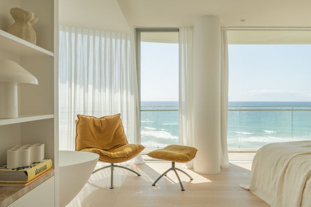 Bright coastal bedroom with direct ocean views through floor-to-ceiling glazing. The room features pale timber flooring, white built-in shelving, sheer curtains, a sculptural white column, and a mustard leather lounge chair with matching ottoman beside a partially visible bed. The overall design is minimal, airy, and refined.