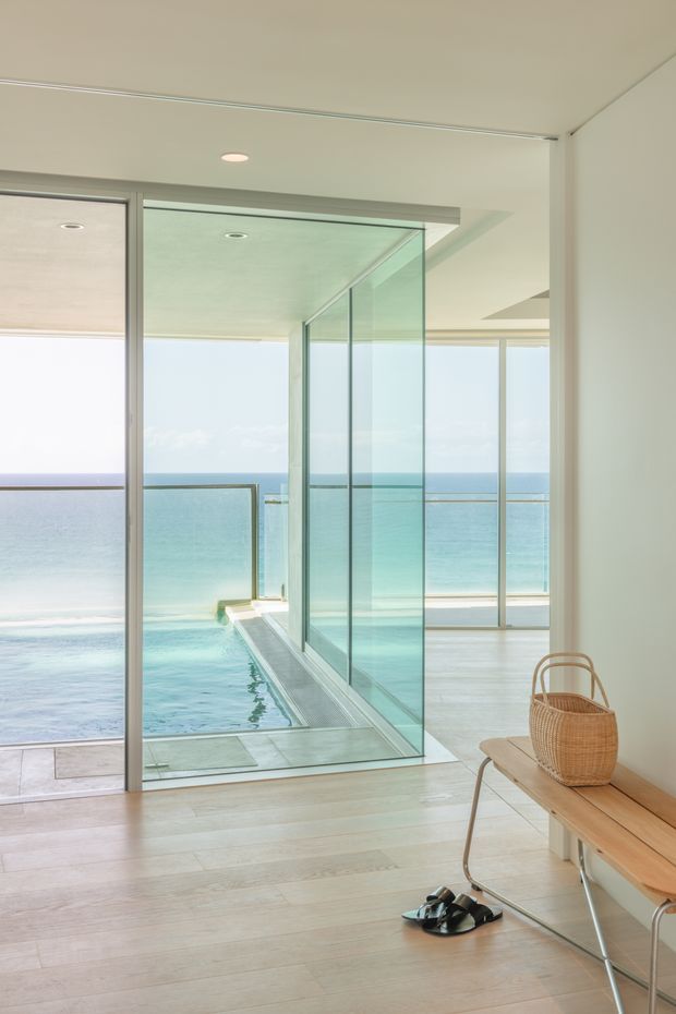 Bright, minimalist coastal interior opening onto a turquoise ocean. The room has pale timber flooring, white walls and ceiling, extensive floor-to-ceiling glazing with slim frames, and a glazed corner or sliding door leading to a narrow poolside or balcony edge. A simple timber bench with a woven basket and sandals sits in the foreground.