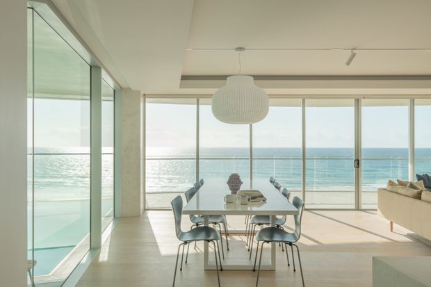 A bright, minimalist open-plan dining and living area with floor-to-ceiling glazing overlooking the ocean and beach. A glass dining table with sculptural dining chairs sits beneath a large ribbed pendant light, while a pale sofa is partially visible at right. Light timber flooring, white surfaces, slim-framed sliding glass doors, and expansive coastal views create an airy, serene interior.
