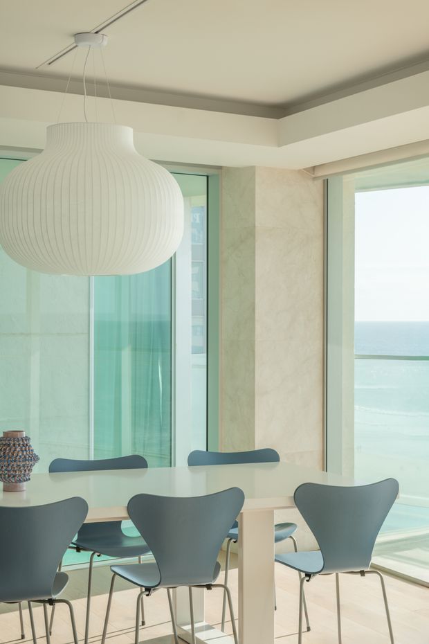 Bright dining area with a pale rectangular dining table and multiple sculptural grey-blue chairs, set beside expansive floor-to-ceiling glazing overlooking the ocean. A large ribbed white pendant light hangs above the table, with pale stone or plaster wall surfaces, light flooring, and a calm coastal palette.