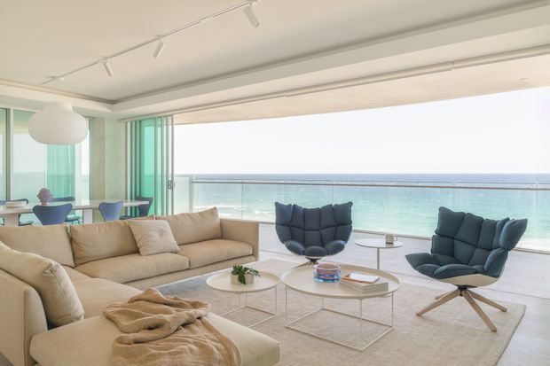 Bright contemporary living room with expansive floor-to-ceiling glazing and sliding glass doors opening onto a balcony with an unobstructed ocean view. The room features a large beige sectional sofa, sculptural dark lounge chairs, round glass coffee tables, a pale area rug, and a linear track-lighting system. A small dining area is visible to the left.
