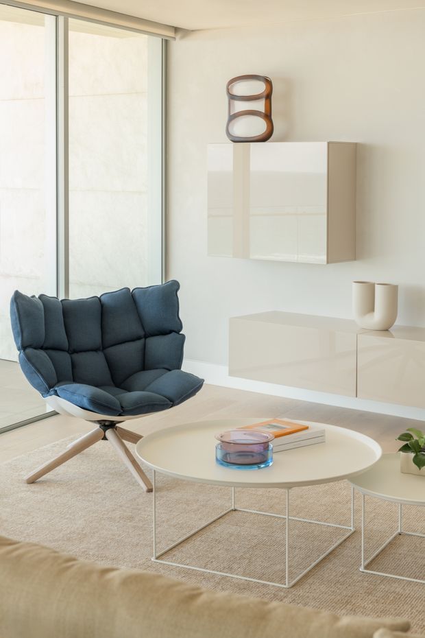 Bright, refined contemporary living room with a sculptural deep blue lounge chair, pale round nesting coffee tables, a low glossy white wall-mounted cabinet, and large floor-to-ceiling glazing. The room uses a soft neutral palette with light timber flooring, textured carpeting, and minimalist decorative objects.