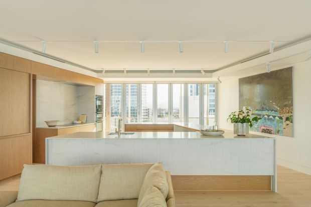 Bright contemporary open-plan apartment interior combining a living area and kitchen. A large white ribbed island dominates the foreground, with warm timber built-in cabinetry and shelving along the left wall, pale timber flooring, white walls and ceiling, and full-height windows overlooking a dense urban skyline. Track lighting and restrained furnishings create a clean, refined atmosphere.