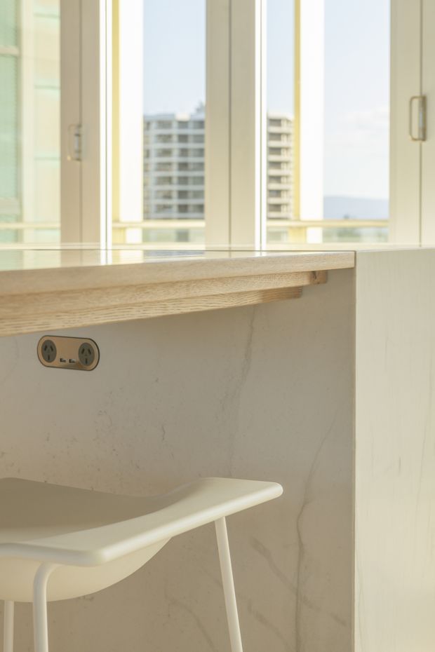 Bright, compact built-in study nook or kitchen-side workspace beside tall white-framed windows. A pale stone or marble-look vertical surface and light timber ledge form the counter, with a double power outlet and a simple white stool. The windows overlook a modern apartment building and distant water or sky, creating an airy contemporary setting.