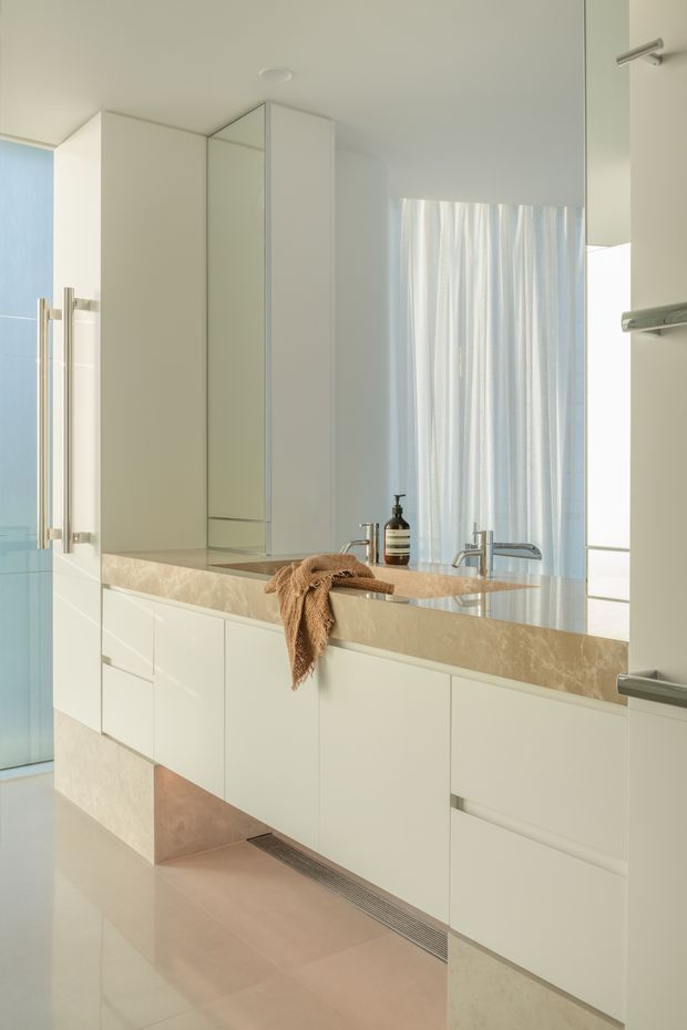 Bright minimalist bathroom featuring a long wall-mounted double vanity with pale cream cabinetry, an integrated beige stone benchtop and backsplash, twin basins, mirrored storage, and brushed metal tapware. A frameless glass shower is visible at left, with soft sheer curtains, glossy light flooring, and warm natural daylight creating a clean, serene atmosphere.