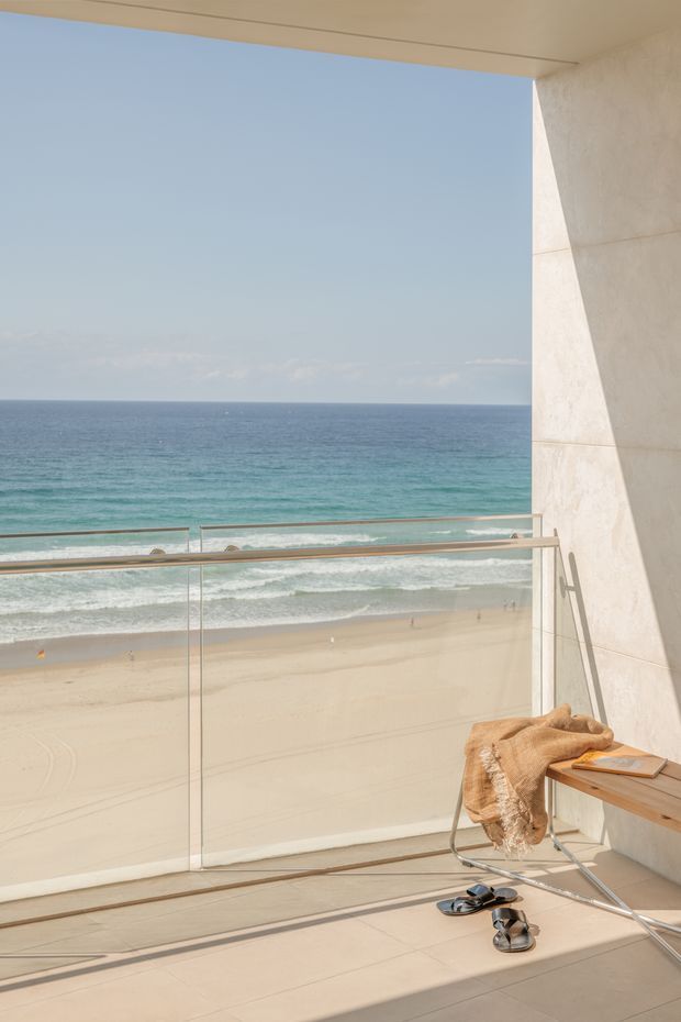 A bright beachfront balcony or terrace overlooking a sandy beach and turquoise ocean. The space features frameless glass balustrades with metal rails, pale large-format floor tiles, a light stone or tiled wall, and a simple timber bench with a folded throw and sandals. The image has a clean, contemporary coastal architectural character with strong indoor-outdoor connection and expansive ocean views.