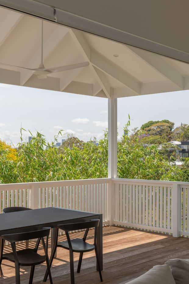 A bright covered outdoor veranda or terrace with a white painted timber roof structure, exposed rafters, ceiling fan, and vertical balustrade. The space has warm timber decking, a dark outdoor dining table with chairs, and views over dense greenery toward neighbouring homes and a distant urban skyline.