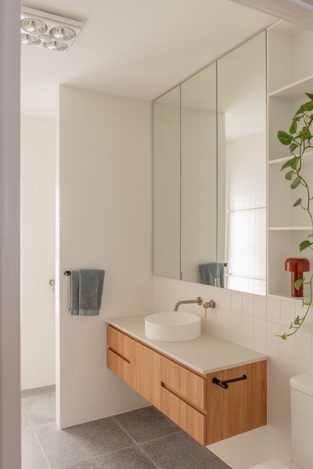 A bright, compact contemporary bathroom with full-height white tiled walls, a large frameless mirrored vanity wall, and a floating light timber vanity with a pale benchtop and round vessel basin. A wall-mounted brushed metal tap, grey towels, open white shelving with a trailing plant, and a modern ceiling light complete the clean, warm interior.