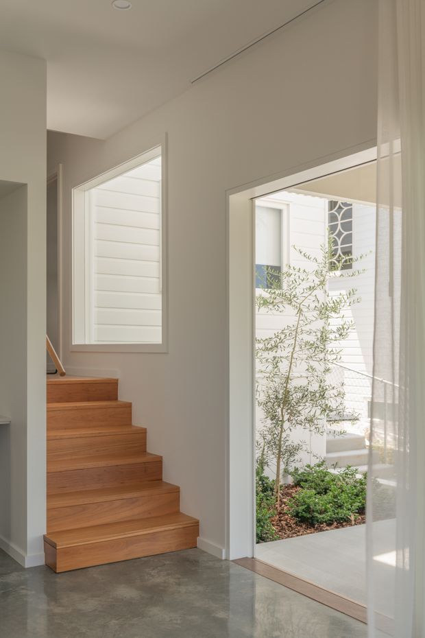 Bright contemporary interior entryway with a straight timber staircase, polished grey concrete flooring, pale painted walls, and full-height glazing overlooking a small planted courtyard. A sheer curtain softens the glazing, while white horizontal exterior cladding and a decorative window are visible beyond.