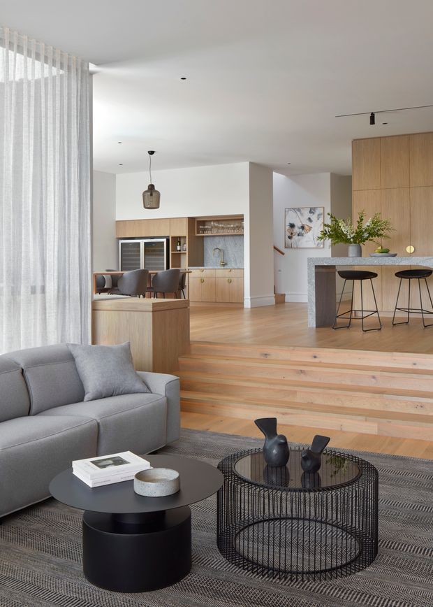 A bright, open-plan contemporary living space viewed from a sunken lounge area. The foreground features a grey upholstered sofa, black round coffee tables, a wire-frame circular side table, and a textured grey rug. Timber steps lead up to a dining area and kitchen, with light timber cabinetry, a dark stone island bench, bar stools, integrated appliances, and a compact drinks or prep station. Sheer floor-to-ceiling curtains, white walls, pale timber flooring, restrained decor, and layered ceiling lighting create an airy, refined interior.