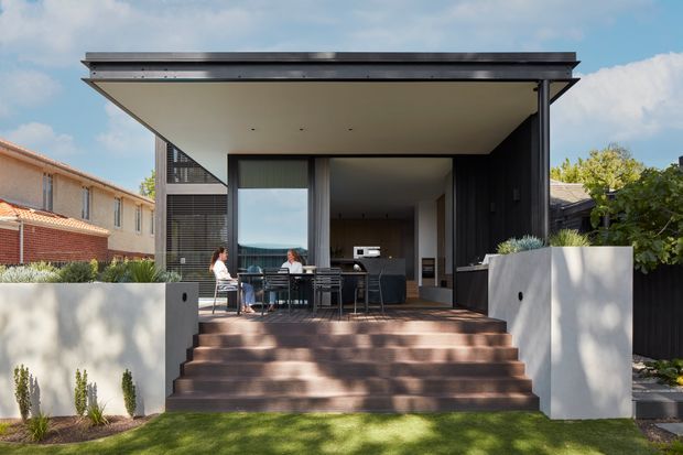 Front-facing exterior view of a contemporary single-storey house extension with a broad flat roof and deep covered terrace. Wide glazed openings connect the sheltered outdoor dining area to a dark interior, while broad steps rise from the lawn between pale rendered planter walls. Dark vertical cladding, slim metal roof edging and neighbouring suburban houses are visible.