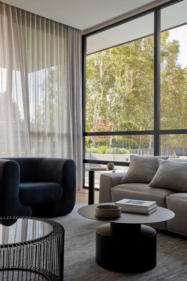 A refined contemporary living room with floor-to-ceiling black-framed glazing overlooking a landscaped outdoor area. The room features sheer and full-height curtains, a deep navy upholstered armchair, a light grey sectional sofa, a round black coffee table, a black cylindrical side table, a wire-frame glass coffee table, and a textured neutral rug. The palette is restrained and sophisticated, with strong natural light and a clear connection to the garden.