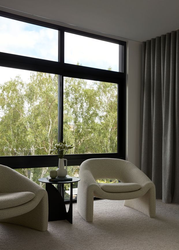 A refined, minimalist sitting area with two sculptural cream boucle lounge chairs beside a small black side table. Full-height black-framed windows overlook leafy trees, while floor-to-ceiling grey curtains and plush neutral carpet create a quiet, soft-toned interior.