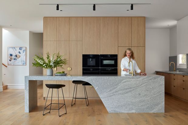 A bright contemporary kitchen with a large light stone island and integrated waterfall end, two black bar stools, warm timber flooring, and a full-height timber-look cabinetry wall incorporating black ovens. Additional timber cabinetry and a dark benchtop continue along the right wall. White walls, track lighting, a vase of greenery, and a small framed artwork complete the clean, refined space.