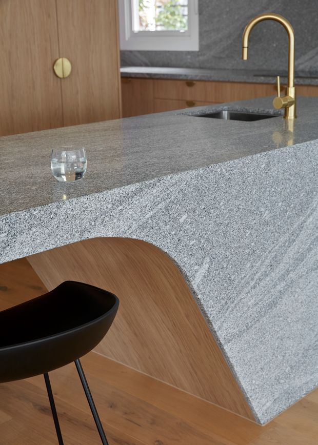 Close-up interior view of a refined kitchen island with a thick grey natural-stone benchtop and waterfall edge, integrated sink, and a curved brass-gold tap. Warm timber cabinetry and timber flooring contrast with the dark stone, while a black sculptural bar stool sits at the island. A small window provides natural light.