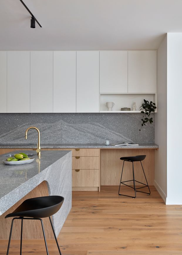 A refined contemporary kitchen with full-height matte white cabinetry, a grey veined stone splashback and island benchtop, warm timber cabinetry and timber flooring, and slim black bar stools. A brass-toned gooseneck tap and black linear track lighting add restrained contrast. The compact layout includes an island with seating and a recessed open display shelf with minimal decor and greenery.