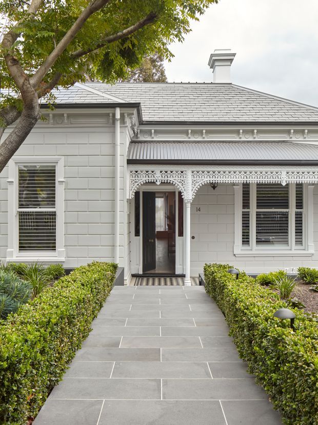 Front exterior view of a refined heritage-style single-storey home, featuring pale painted masonry walls, ornate decorative timber lacework across the verandah, classical cornices and mouldings, a hipped grey tiled roof with chimney, timber-framed windows with horizontal blinds, and a formal paved path bordered by clipped hedges and landscaped planting.