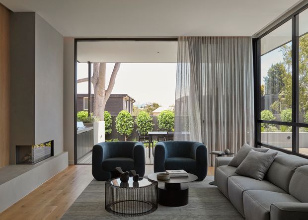 A refined contemporary living room with warm timber flooring, a minimalist linear fireplace, full-height glazing and sheer curtains opening onto a planted outdoor terrace. Two dark lounge chairs and a large light-grey sofa surround low round coffee tables on a textured rug. The room is bright, calm, and connected to the exterior through expansive windows and sliding doors.