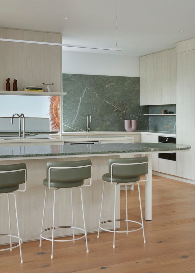 A bright contemporary kitchen centred on a large island with a green stone benchtop and seating for four. Pale timber cabinetry extends across the walls, with a matching pale timber island base, integrated appliances, a linear pendant light, and a dramatic green veined stone splashback. The kitchen opens toward a secondary area visible through a pass-through, while muted olive-green bar stools and a pink ceramic vessel add restrained colour accents.