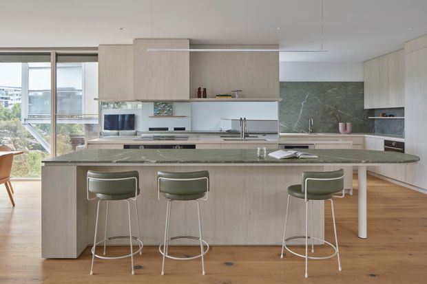 Bright contemporary kitchen with a large island and three upholstered bar stools, pale timber cabinetry, a dark stone island benchtop, integrated appliances, and a green-grey stone splashback. Floor-to-ceiling glazing on the left provides views toward the surrounding urban area. The space has a clean, refined, open-plan feel.