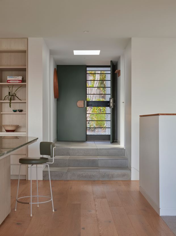 A bright, modern residential entryway viewed from inside. Wide timber floorboards lead to a raised entry landing with broad grey tiled steps and a dark contemporary front door incorporating a black metal grid and glazed panels. Built-in light timber shelving and cabinetry line the left side, with a small bar-height stool beside a stone or composite counter. A white stair or wall partition with a timber cap is visible on the right. A rectangular skylight or ceiling light brightens the corridor.